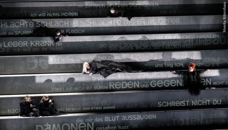 performers on set consisting of movable steps, stairs and drawers with German text projected on them