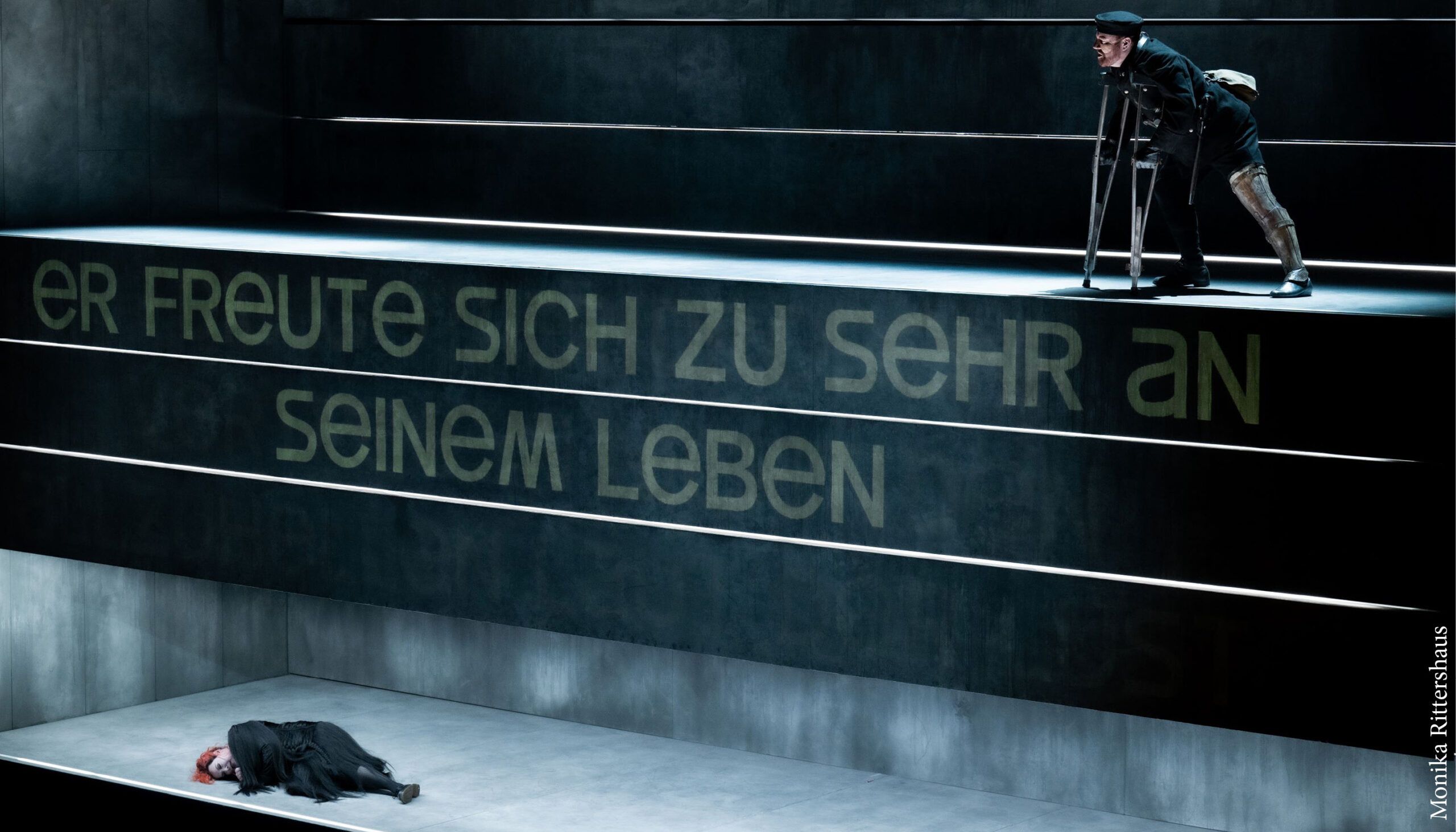 two performers on set consisting of movable steps, stairs and drawers with German text projected on them