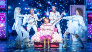 Musical theatre scene showing performers in white ABBA-inspired costumes surrounding a woman in pink dress sitting on a bed with heart pattern duvet.