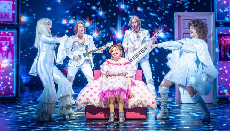Musical theatre scene showing performers in white ABBA-inspired costumes surrounding a woman in pink dress sitting on a bed with heart pattern duvet.