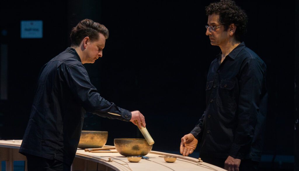 Two men in dark clothing performing with singing bowls on wooden table in dimly lit performance space