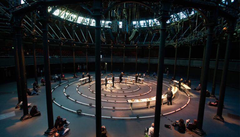 Large circular performance space with singing bowls arranged in spiral pattern, audience seated around perimeter watching performers
