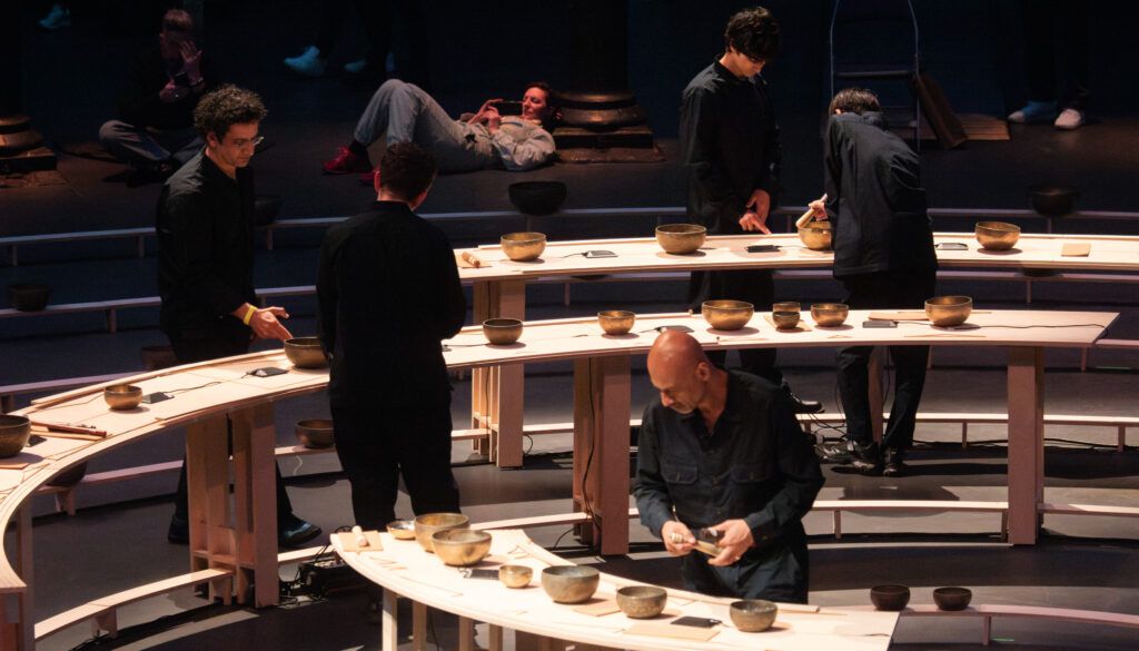 Multiple performers in black clothing playing singing bowls at wooden tables in performance space with tiered seating