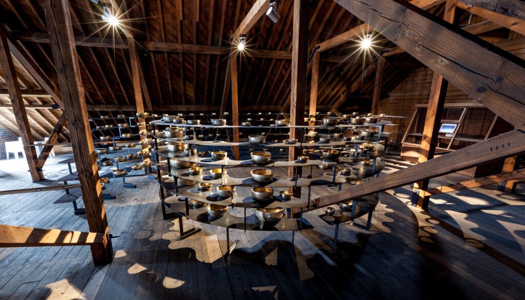Space filled with hundreds of singing bowls arranged on metal stands under exposed beam ceiling