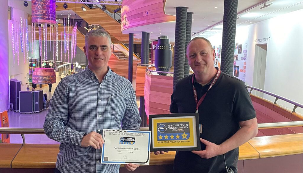 Two men holding security awards in modern building lobby with colourful lighting and multi-level architecture
