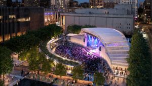Nighttime concert at outdoor amphitheatre with curved translucent roof. Large audience gathered before illuminated stage with city skyline visible behind performers.