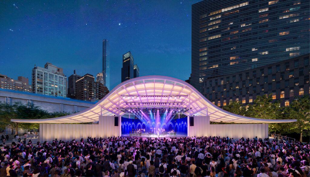 Illuminated outdoor performance pavilion with distinctive curved shell roof glowing purple under night sky. Dense crowd watches concert against backdrop of city buildings.