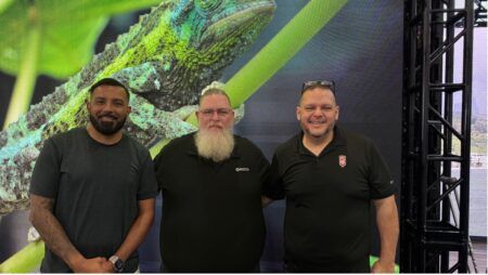 Three men posing together in front of large display showing an iguana