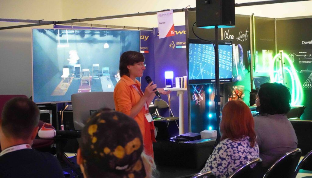 Woman in orange shirt presenting to seated audience with screens and lighting displays at trade show