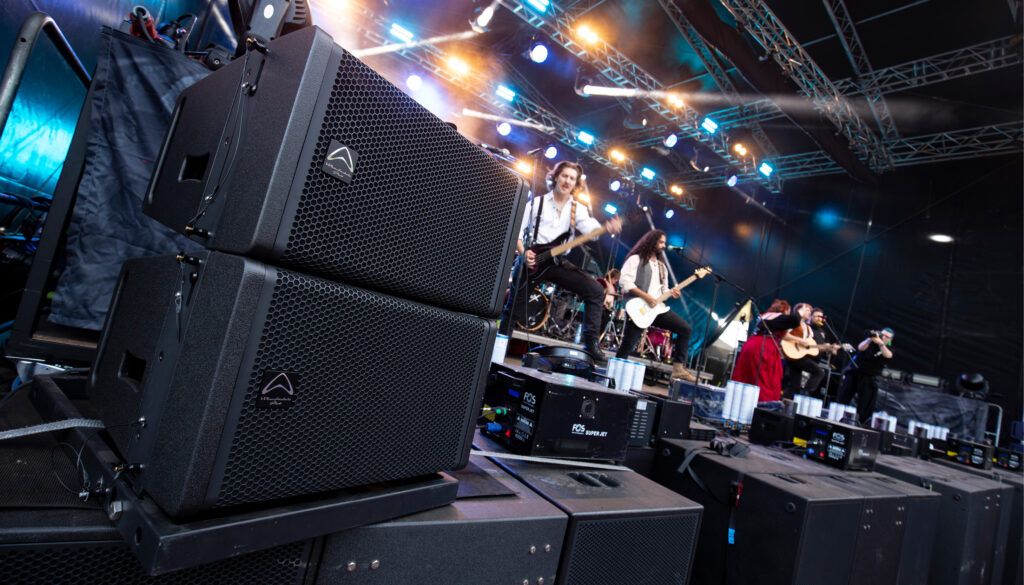 Professional sound equipment stacked at side of outdoor concert stage with band performing under colourful lights
