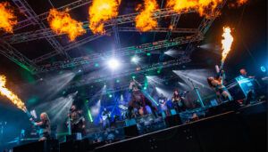 Dramatic concert scene with performers, pyrotechnics, flame effects, and green stage lighting creating spectacle