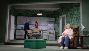 Theatre stage set with man and woman talking in kitchen, and older woman in armchair