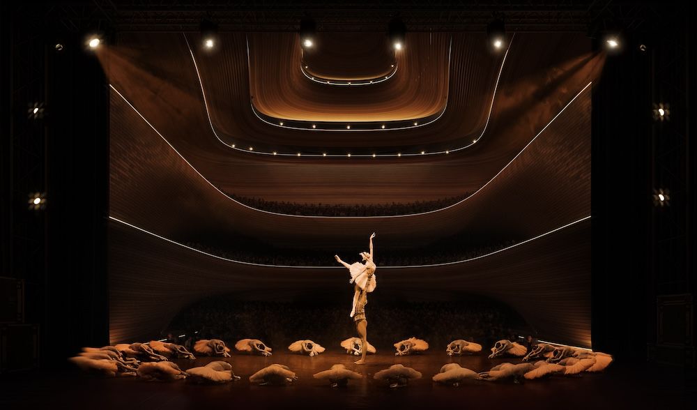 Interior performance view of The Opera & Ballet Theatre of Kosovo showing concentric curved timber balconies rising above a stage where a duo of ballet dancers performs, encircled by a group of ballet dancers in swan pose on the stage floor.