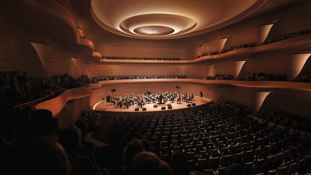 Interior view of the Opera & Ballet Theatre of Kosovo's main auditorium showing curved timber-clad balconies and walls surrounding a central orchestra stage, with a full audience in attendance and musicians performing.