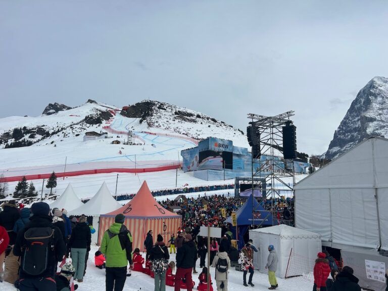 Panoramic view of a Lauberhorn outdoor spectator area, showing the snow-covered course, a large LED scoreboard screen, rigged speaker stacks, colourful hospitality tents and a mountain backdrop.