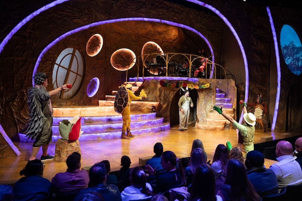 Performers in animal costumes on a curved, earth-toned stage set with circular openings and purple accent lighting, watched by a full seated audience.
