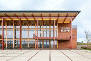 Exterior view of the redeveloped Theatr Clwyd building showing the new glazed timber foyer structure, red columns, brick facade and illuminated venue signage