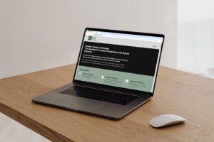 A laptop on a wooden desk displaying the System Design Exchange homepage, introducing the SDE standard for noise predictions with sound systems.