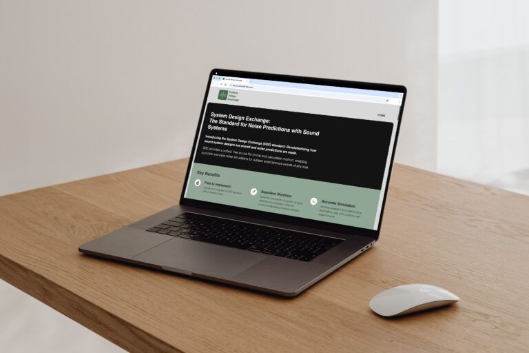 A laptop on a wooden desk displaying the System Design Exchange homepage, introducing the SDE standard for noise predictions with sound systems.