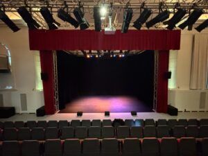 Interior view of the Noorderkerkzaal theatre, showing rows of seating, a stage framed by red curtains, speakers and overhead lighting rigs.