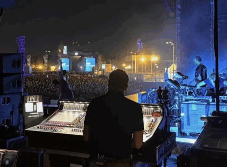 Photograph of a darkened arena front of house mixing position with illuminated audio consoles and outboard equipment racks during a live concert