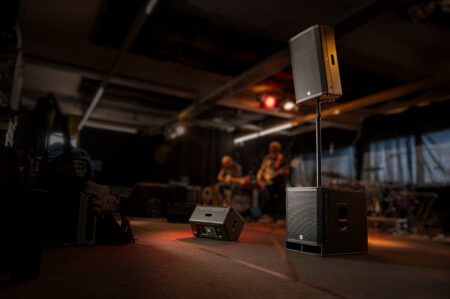 Portable PA speaker system on subwoofer pole mount in dimly lit rehearsal room with band playing in background