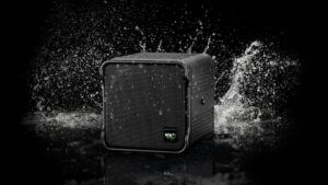 A black cube-format loudspeaker bearing the KV2 Audio logo surrounded by water splashing against it on a dark reflective surface.