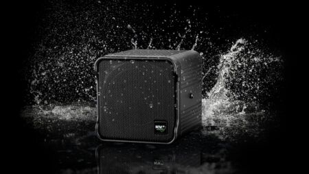 A black cube-format loudspeaker bearing the KV2 Audio logo surrounded by water splashing against it on a dark reflective surface.