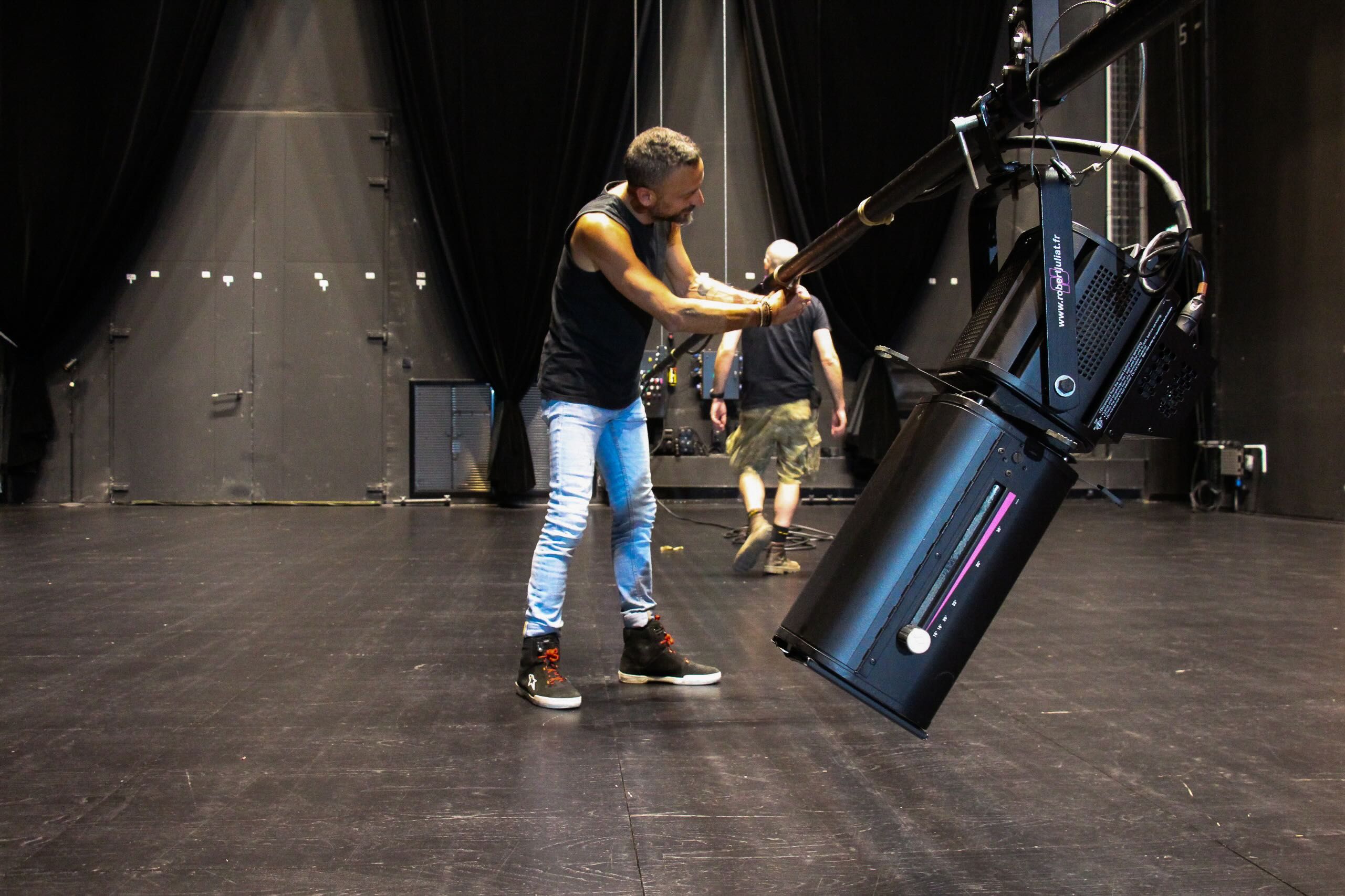 A technician adjusting a black Robert Juliat profile spotlight rigged on a bar on a bare theatre stage