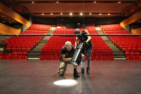 Robert Juliat retrofit brings LED to La Théâtre de Laval Two technicians fitting a black profile spotlight to a lighting bar on a theatre stage, with an empty red-seated auditorium behind them