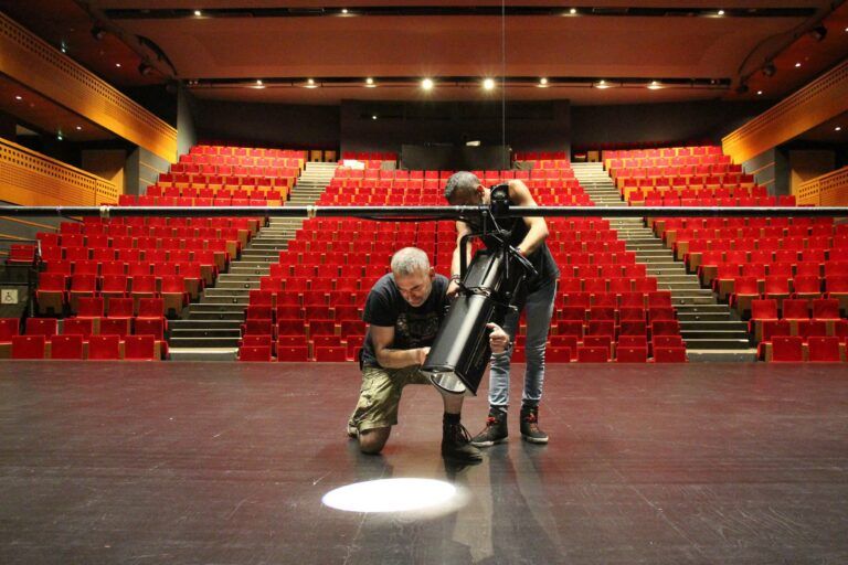Two technicians fitting a black profile spotlight to a lighting bar on a theatre stage, with an empty red-seated auditorium behind them