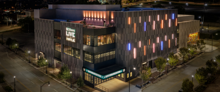 A large modern concert venue lit at night, its dark textured facade marked with vertical strips of orange and blue light and illuminated ‘Citizens Live at The Wylie’ signage, on a street corner in Pittsburgh’s Hill District.
