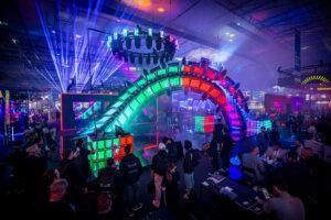 A trade show stand with a large arch of illuminated red and green LED cube panels beneath a circular lighting rig emitting beams of light, as visitors photograph the display and meet at nearby tables.
