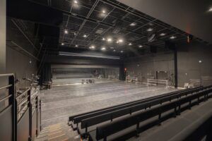 Black-box studio theatre with raked bench seating, grey dance floor, ballet barres, mirrored back wall and grid ceiling.