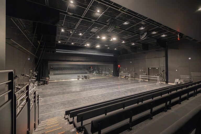 Black-box studio theatre with raked bench seating, grey dance floor, ballet barres, mirrored back wall and grid ceiling.