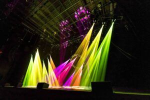Coloured beams of green, yellow, orange and magenta light from fixtures across a stage, with a circular truss in the centre.