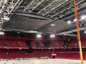 Interior of Johan Cruijff Arena with red seating, showing large black acoustic baffle panels suspended under the steel roof trusses, black drapes hung over sections of the stands and access equipment on the covered pitch